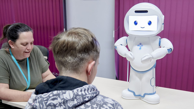 Student and teaching sitting down at a desk in front of a robot that teaches students social skills.