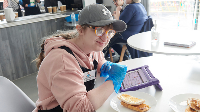 Woman wearing glasses and a cap sitting down at a table eating toast. She has a tablet next to her.