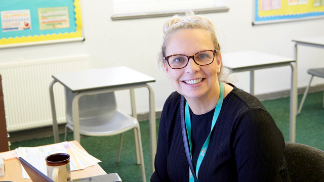 Woman sitting down at her desk in a classroom. She is smiling wide at the camera. 