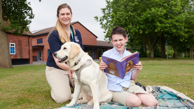 Young boy holding a story book about a dog sitting down on a blanket outside. Next to him there is an adult woman wearing a teacher uniform holding a yellow labrador. 