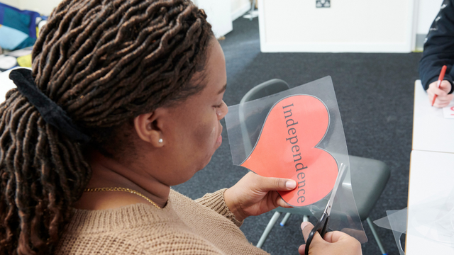 Adult woman holding a paper heart that has "independence" written on it. She is cutting around the heart to make a collage. 