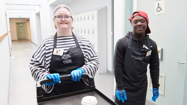 Young man and woman, both with down syndrome, wearing aprons and hair nets. The woman is pushing a trolley with a coffee cup on it. They are both wearing badges that read "barista"  