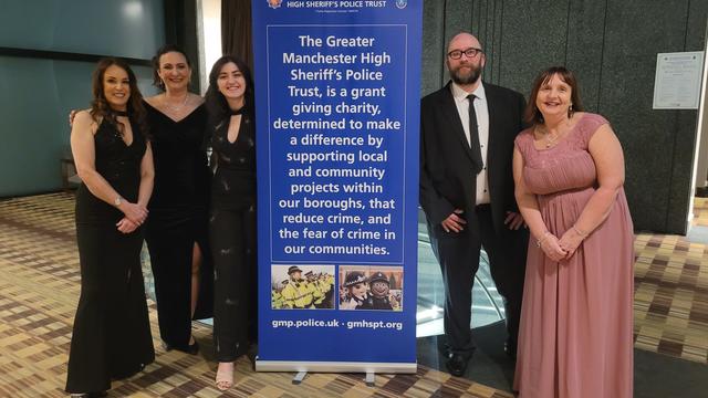 5 members of staff dressed in elegant clothes standing next to a banner that reads "The greater manchester high sheriff's police trust is a grant giving charity determined to make a difference by supporting local and community projects within our boroughs, that reduce crime and the fear of crime in our communities" 