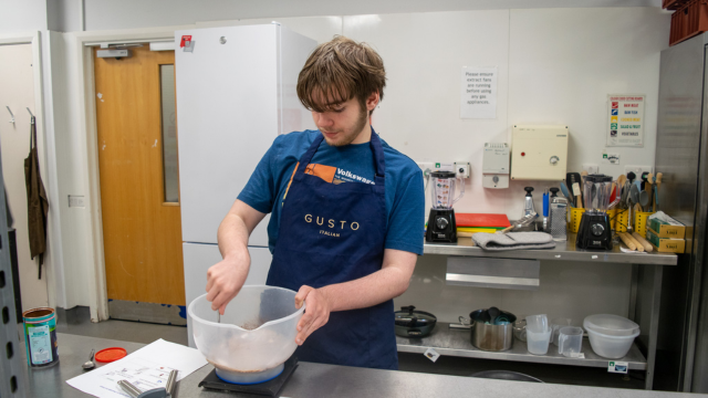 teen boy mixing batter in a bowl with a whisk. he is wearing an apron that says "Gusto" and is working in an industrial style kitchen. 