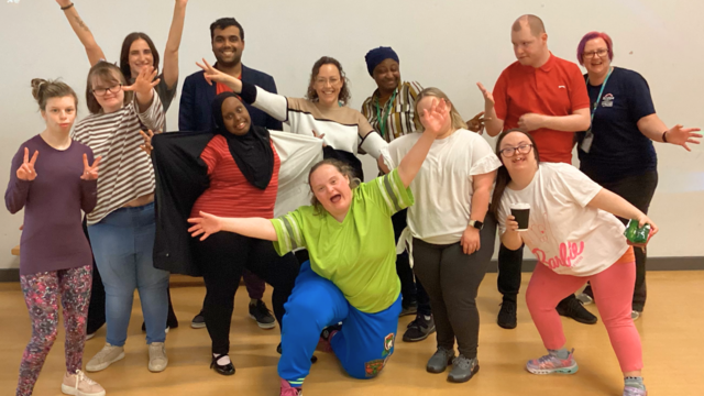 A group of students standing together smiling and waving at the camera. Hannah the Zumber teachers is knelt down in the middle of the image with her arms in the air.